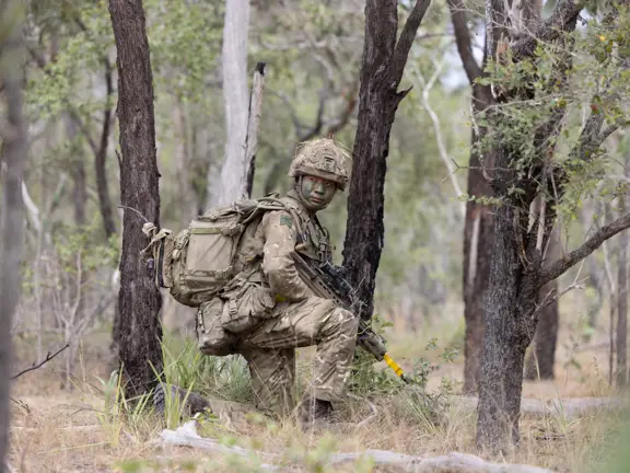 A person in military camouflage, including face paint and gear, is positioned in a forested area, holding a rifle.