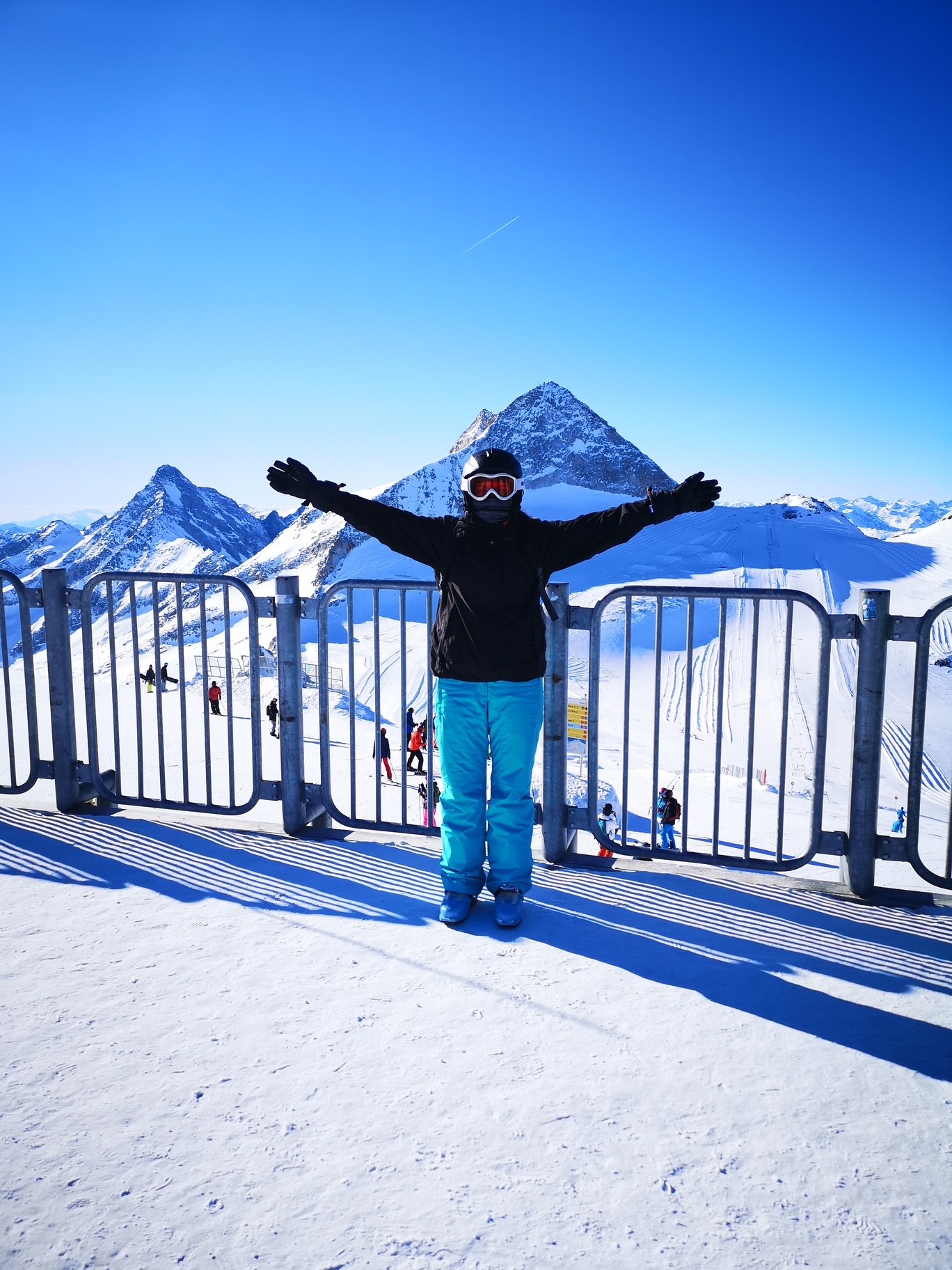 A person in winter gear poses on a snowy mountaintop with arms outstretched, against a bright blue sky and rugged peaks. The atmosphere is joyful and adventurous.