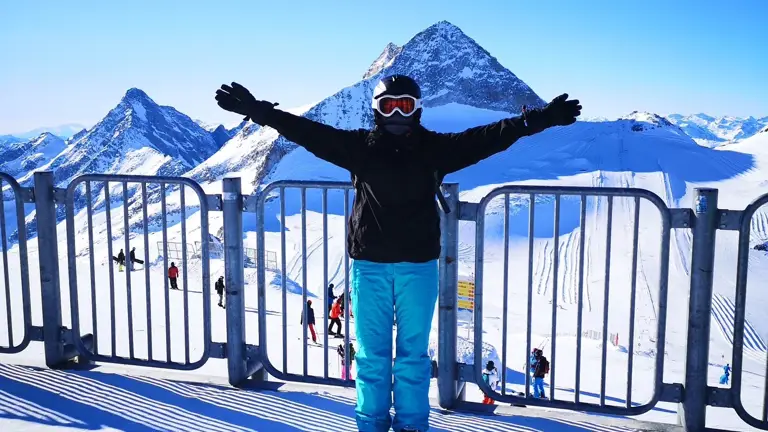 A person in winter gear poses on a snowy mountaintop with arms outstretched, against a bright blue sky and rugged peaks. The atmosphere is joyful and adventurous.