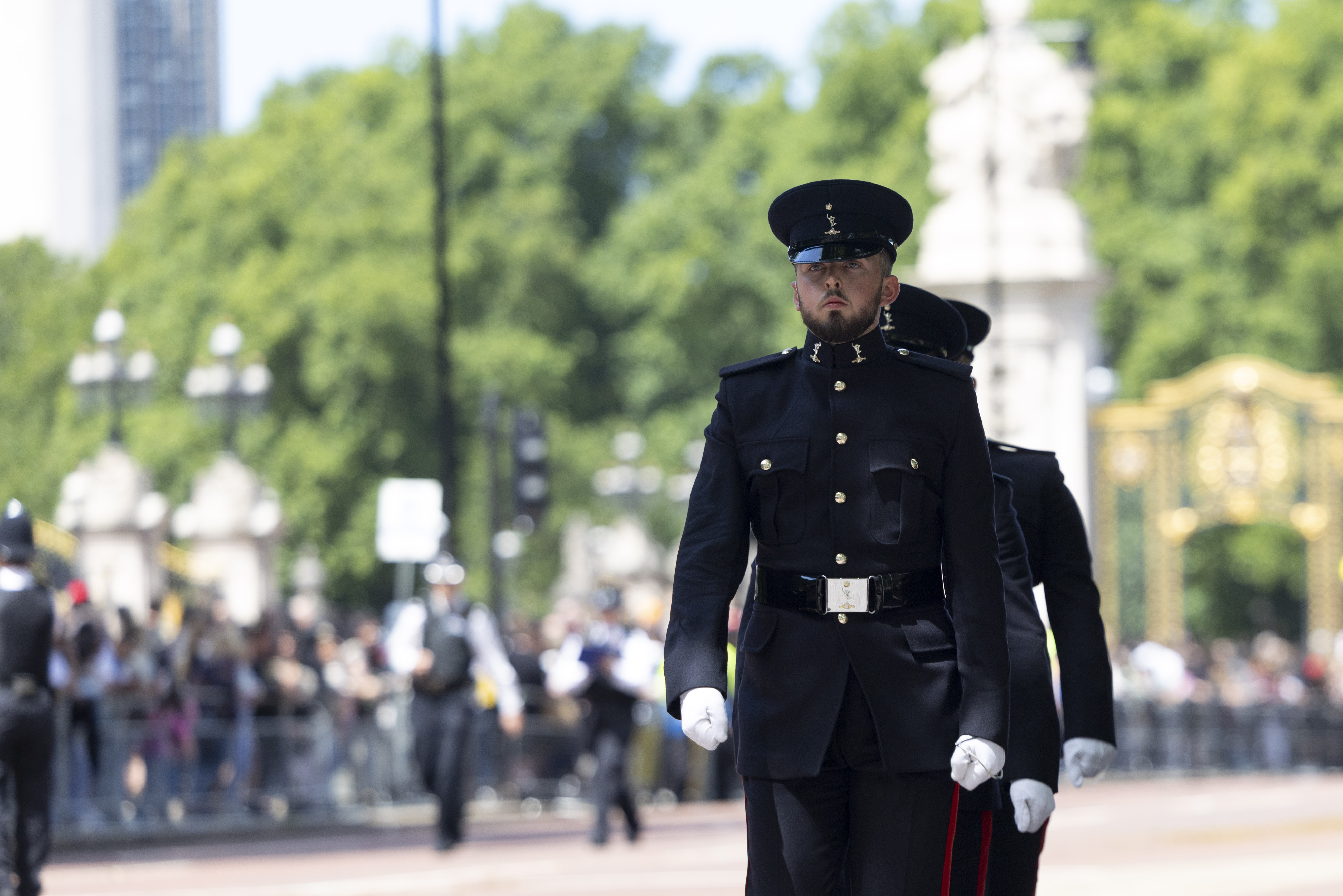 Soldiers in dark uniforms march in formation at a ceremonial event, with blurred crowds and greenery in the background. The scene conveys formality.