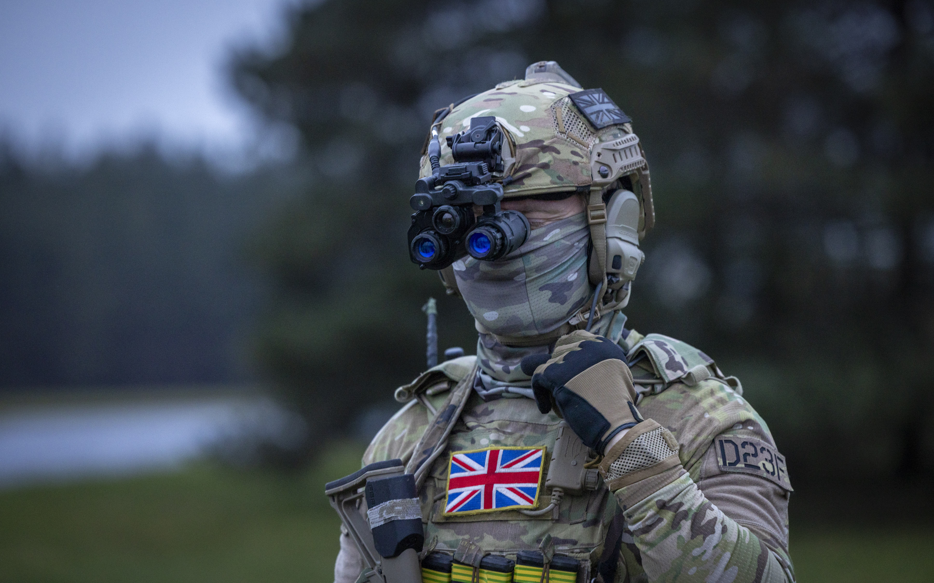 Soldier in full camouflage gear wearing night vision goggles and a UK flag patch on the chest, standing outdoors.