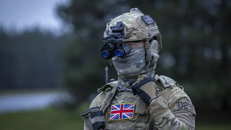 Soldier in full camouflage gear wearing night vision goggles and a UK flag patch on the chest, standing outdoors.