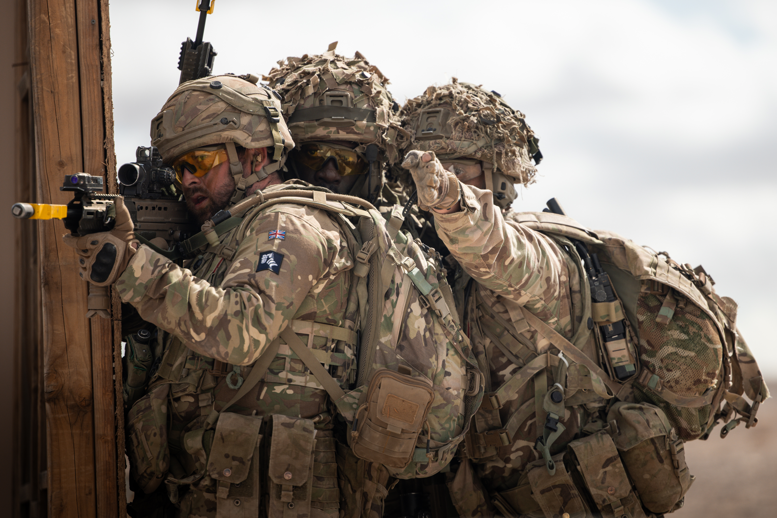 A soldier stands aiming his rifle while two soldiers behind him point at a target.