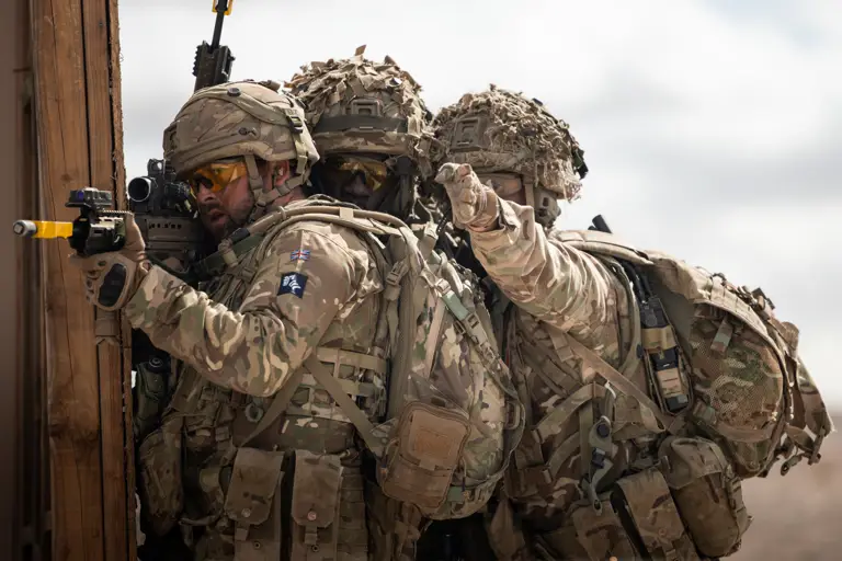 A soldier stands aiming his rifle while two soldiers behind him point at a target.