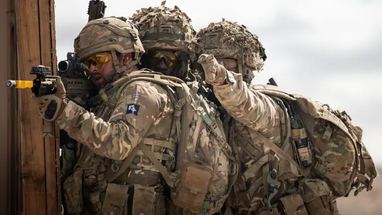 A soldier stands aiming his rifle while two soldiers behind him point at a target.
