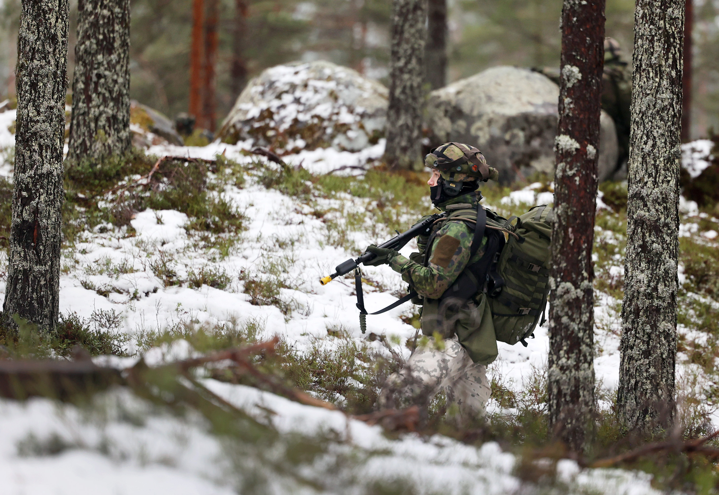 A soldier in camouflage gear and helmet walks through a snowy, forested area. They carry a rifle and backpack, blending with the quiet, winter landscape.