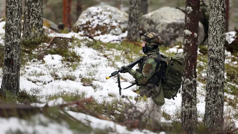 A soldier in camouflage gear and helmet walks through a snowy, forested area. They carry a rifle and backpack, blending with the quiet, winter landscape.