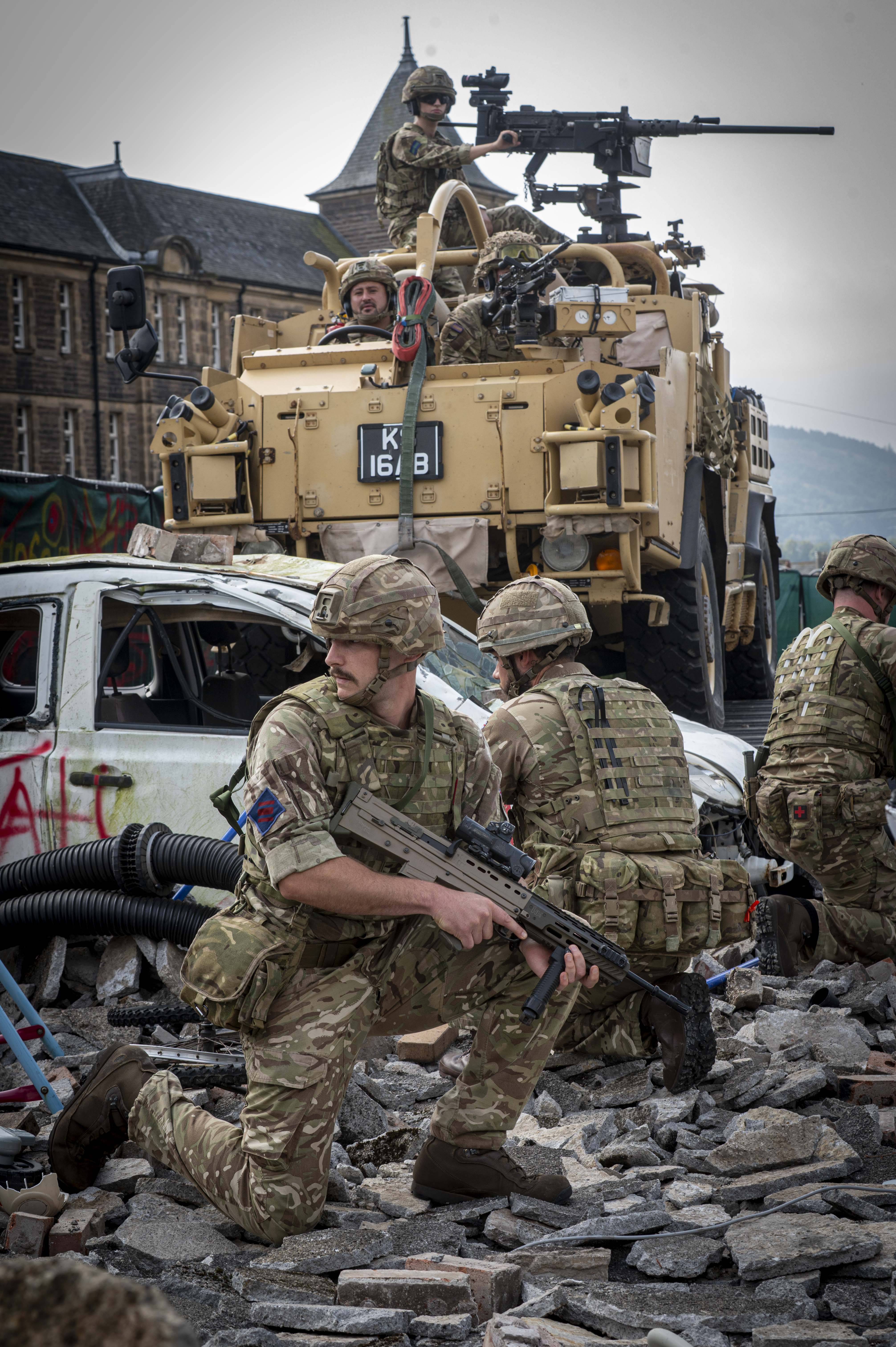 Soldiers in camouflage gear take cover among rubble and debris near a military vehicle equipped with heavy weaponry.