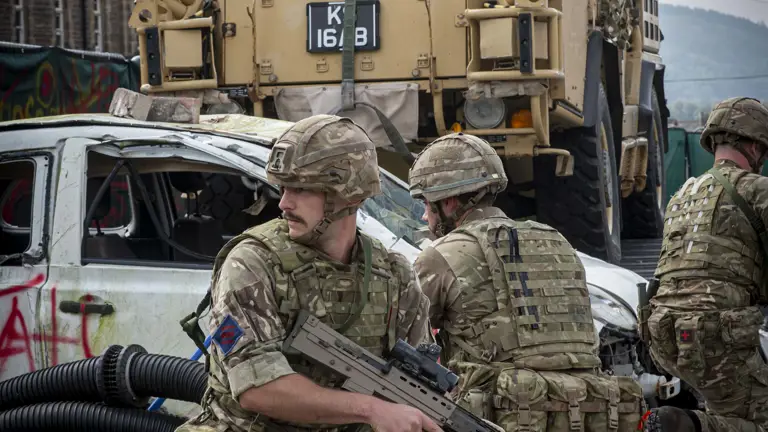 Soldiers in camouflage gear take cover among rubble and debris near a military vehicle equipped with heavy weaponry.