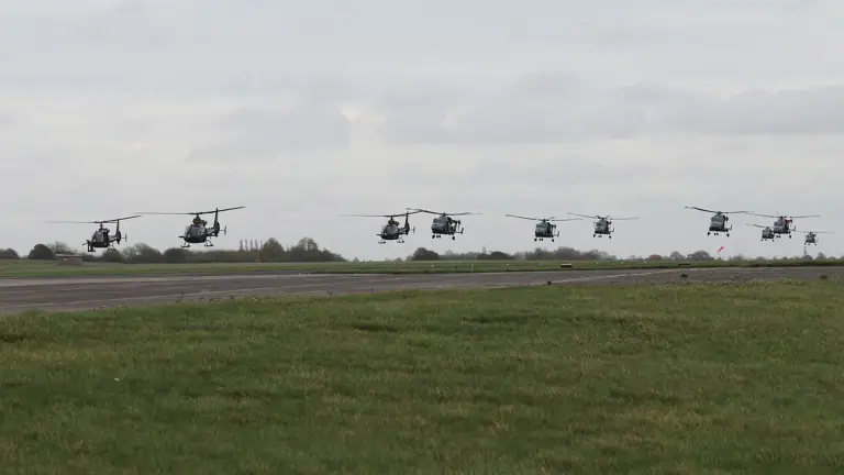 Multiple military helicopters flying low in a staggered formation over a grassy airfield under a cloudy sky.
