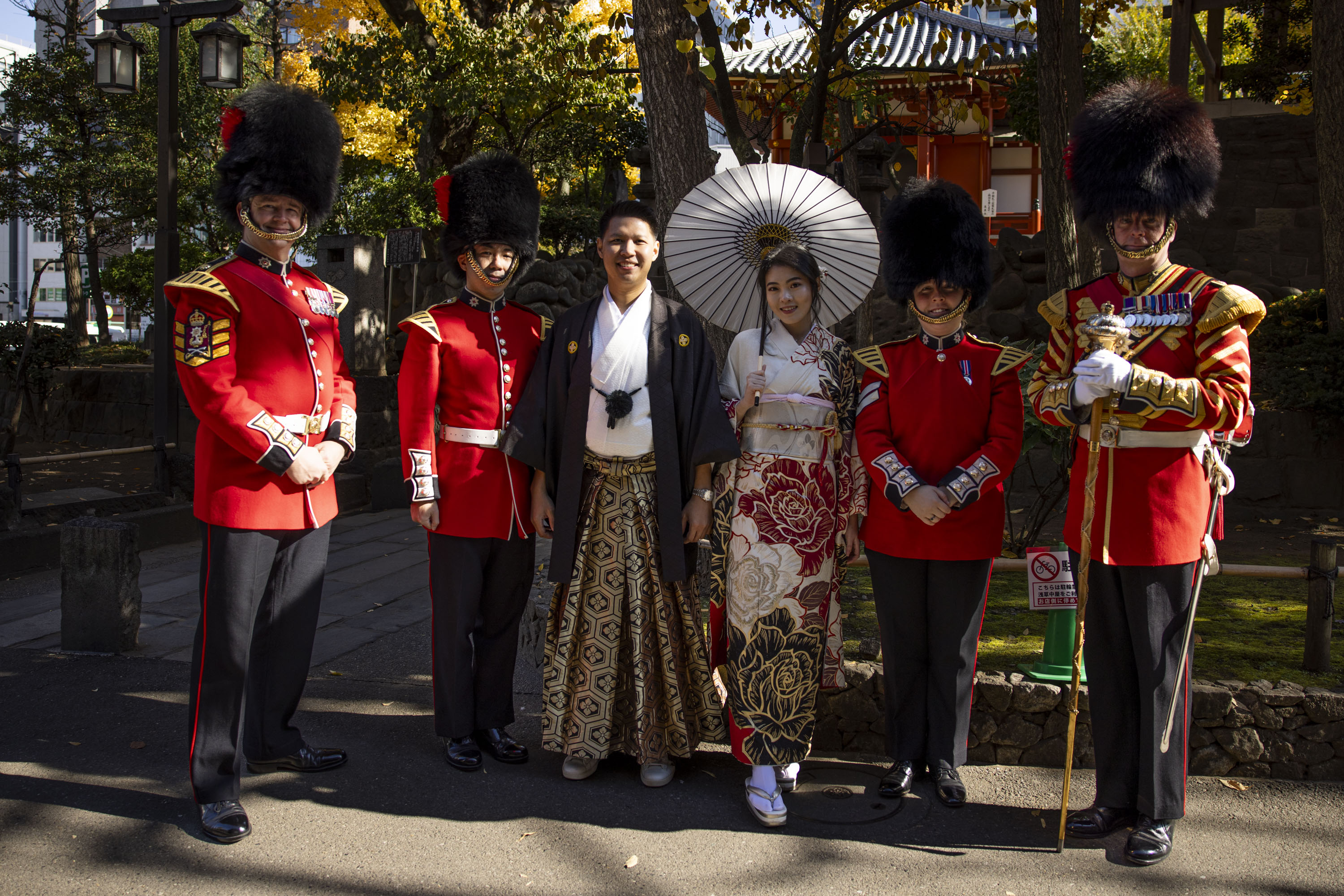 Four soldiers in red tunics and bearskin hats are pictured with two people in traditional Japanese dress. 