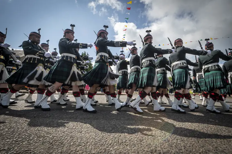 Men wearing black tunics and green and blue kilts march past. They are carrying rifles.