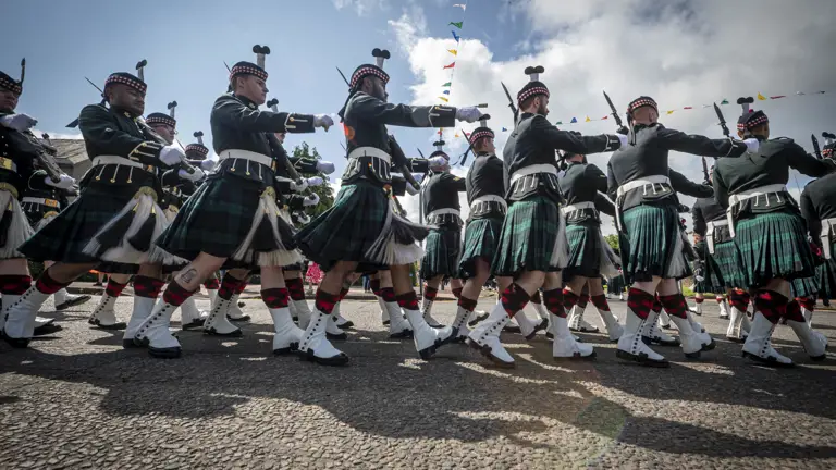 Men wearing black tunics and green and blue kilts march past. They are carrying rifles.