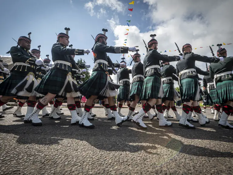 Men wearing black tunics and green and blue kilts march past. They are carrying rifles.