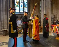 Two soldiers stood in a cathedral wearing ceremonial uniform , one is handing a large flag known as colours to a lady wearing a church robe. A man stood behind her wears a black and red church robe.