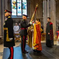 Two soldiers stood in a cathedral wearing ceremonial uniform , one is handing a large flag known as colours to a lady wearing a church robe. A man stood behind her wears a black and red church robe.