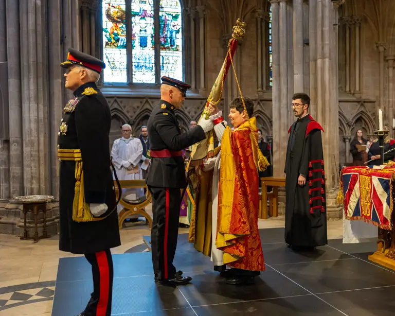 Two soldiers stood in a cathedral wearing ceremonial uniform , one is handing a large flag known as colours to a lady wearing a church robe. A man stood behind her wears a black and red church robe.