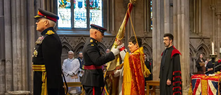 Two soldiers stood in a cathedral wearing ceremonial uniform , one is handing a large flag known as colours to a lady wearing a church robe. A man stood behind her wears a black and red church robe.