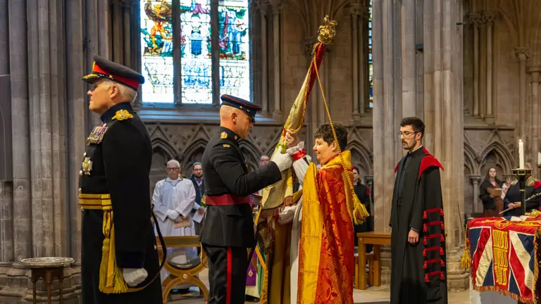 Two soldiers stood in a cathedral wearing ceremonial uniform , one is handing a large flag known as colours to a lady wearing a church robe. A man stood behind her wears a black and red church robe.