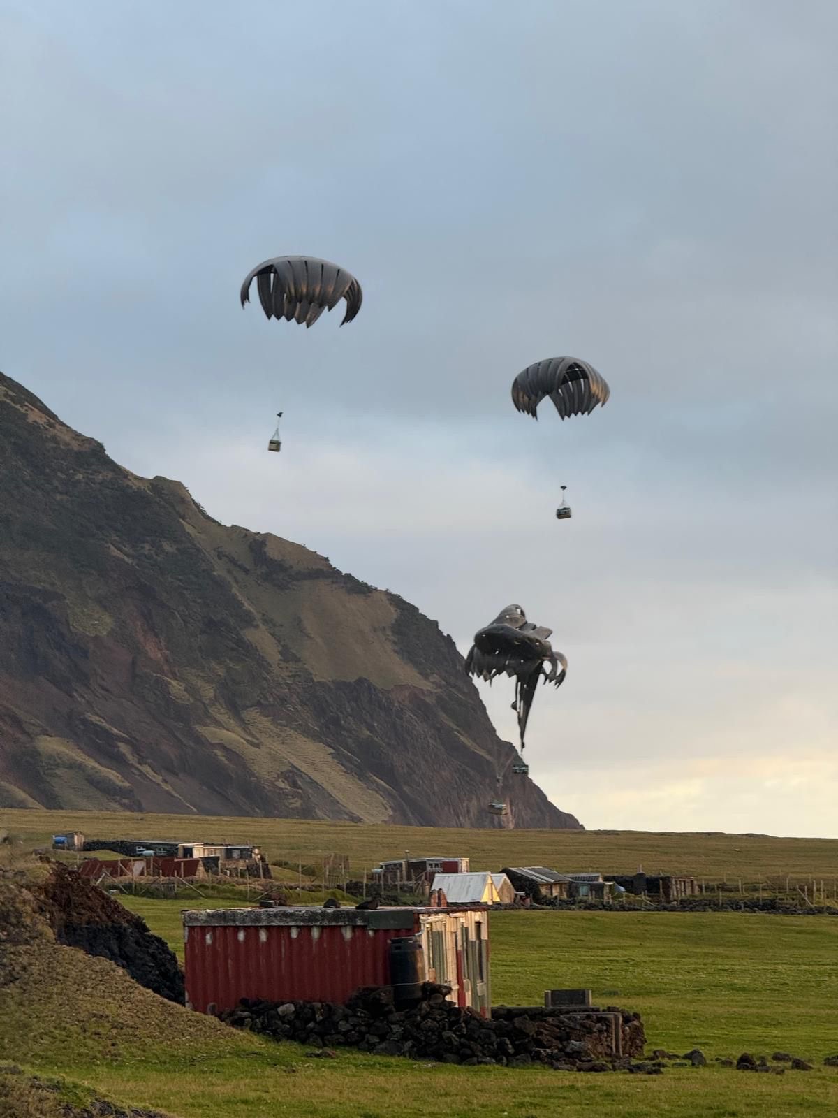 Three parachutes descend over a rural landscape with green fields, mountains, and small buildings. The scene feels serene and adventurous.