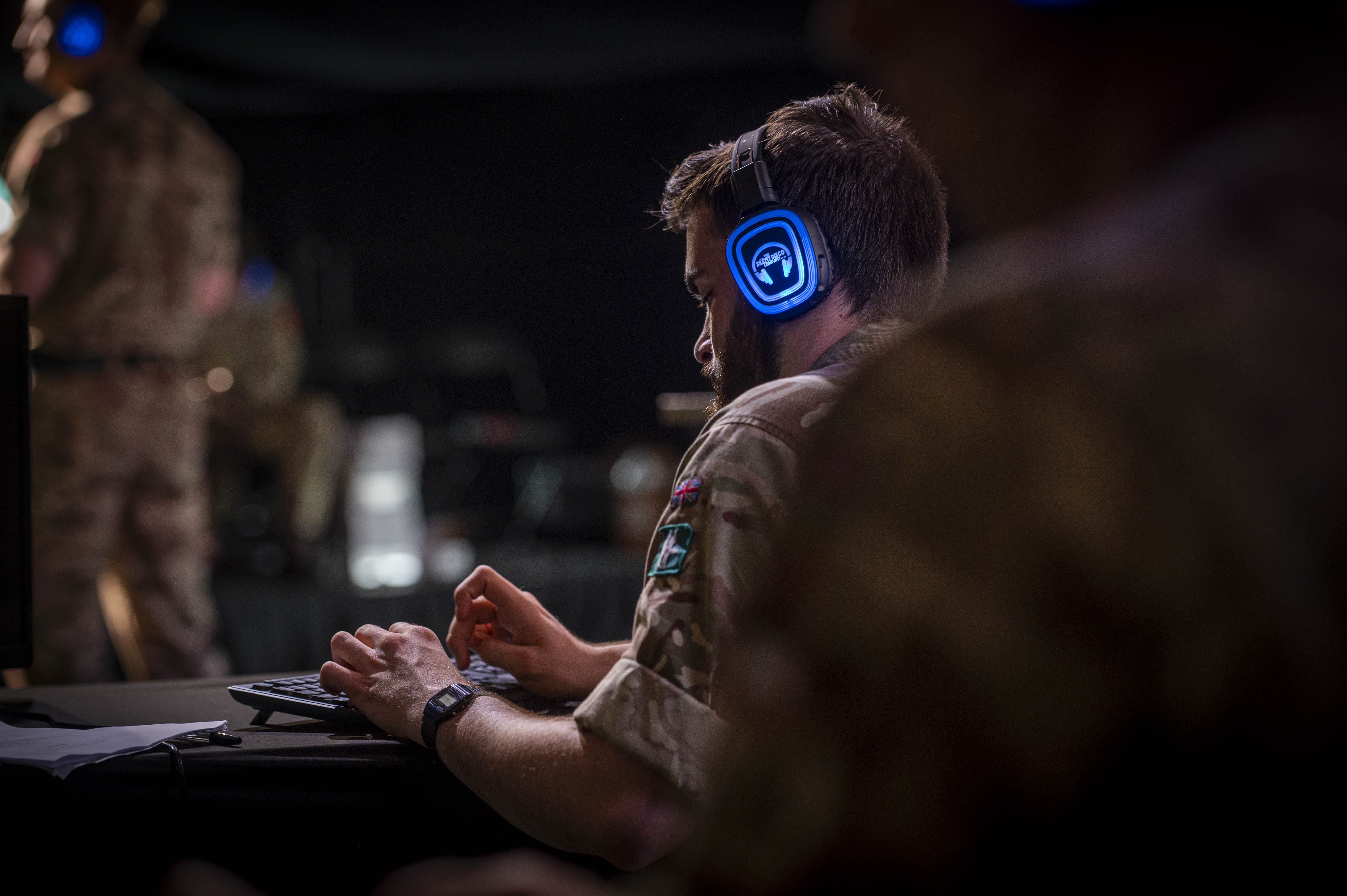 Soldier in camouflage uniform wearing blue headphones and typing on a keyboard in a dimly lit room.