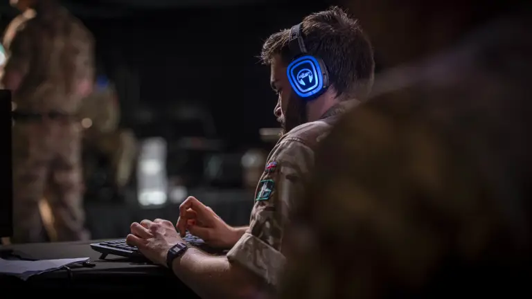 Soldier in camouflage uniform wearing blue headphones and typing on a keyboard in a dimly lit room.