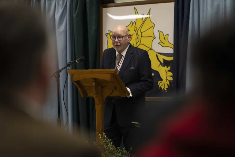 A man in a suit speaks at a podium with a microphone, against a backdrop featuring a yellow dragon emblem. The atmosphere is formal and attentive.