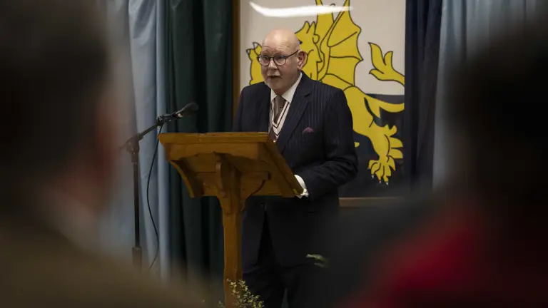A man in a suit speaks at a podium with a microphone, against a backdrop featuring a yellow dragon emblem. The atmosphere is formal and attentive.