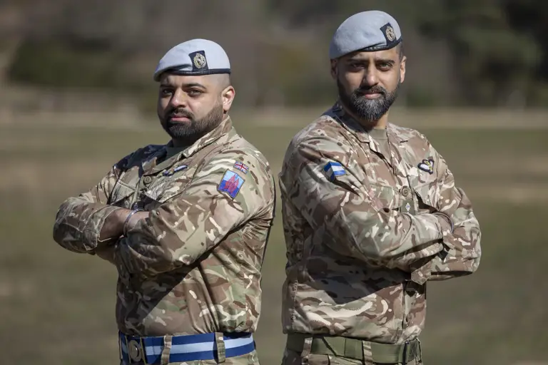 Two soldiers in their camouflage unifrom with their arms folded and light blue berets on their heads stood next to each other outside.