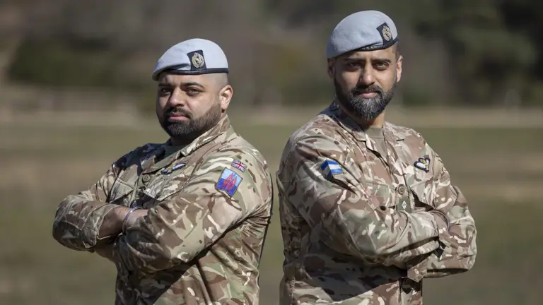 Two soldiers in their camouflage unifrom with their arms folded and light blue berets on their heads stood next to each other outside.