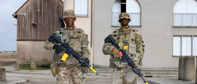 Two soldiers in camouflage uniforms stand next to each other holding rifles.