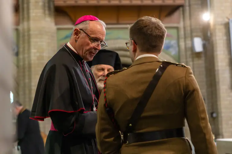 A clergyman in black and red robes speaks with a soldier in a brown uniform inside a church with brick walls.