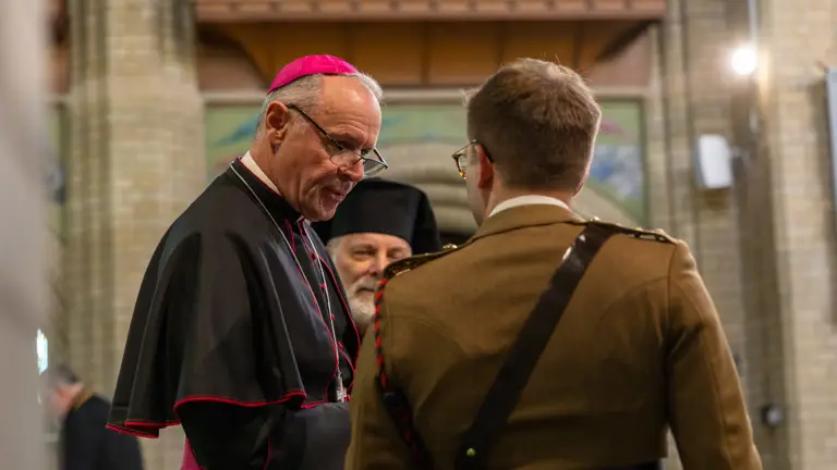 A clergyman in black and red robes speaks with a soldier in a brown uniform inside a church with brick walls.