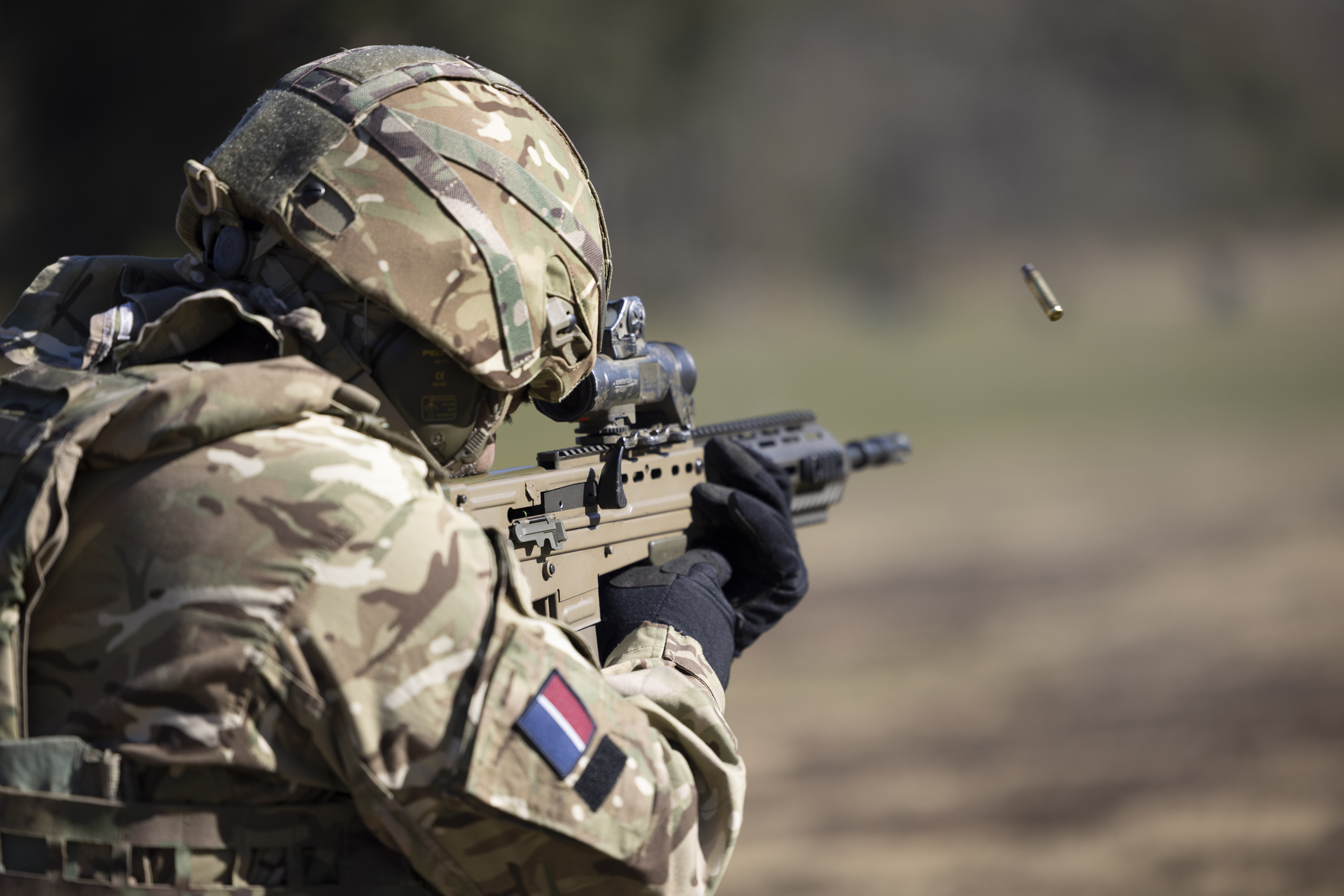 A soldier in British Army uniform is pictured looking down a scope and firing.