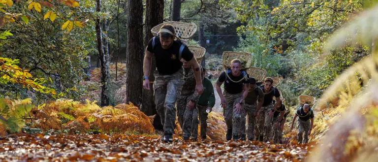 Group of soldiers carrying large backpacks climbing a forest trail covered in autumn leaves under tall trees.