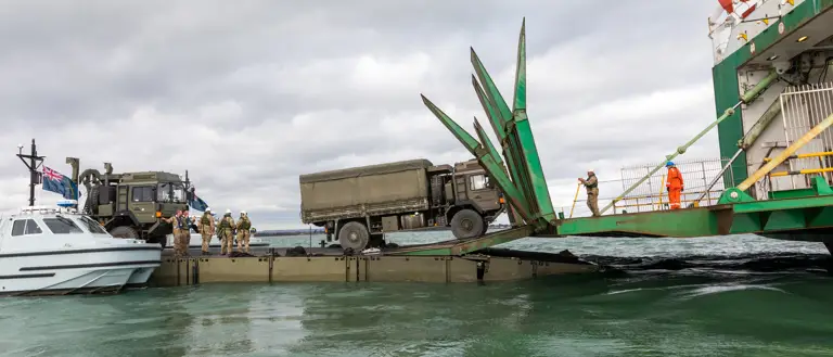 A military truck drives onto a ship via a MEXEFLOTE which is a landing platform which floats on the water.
