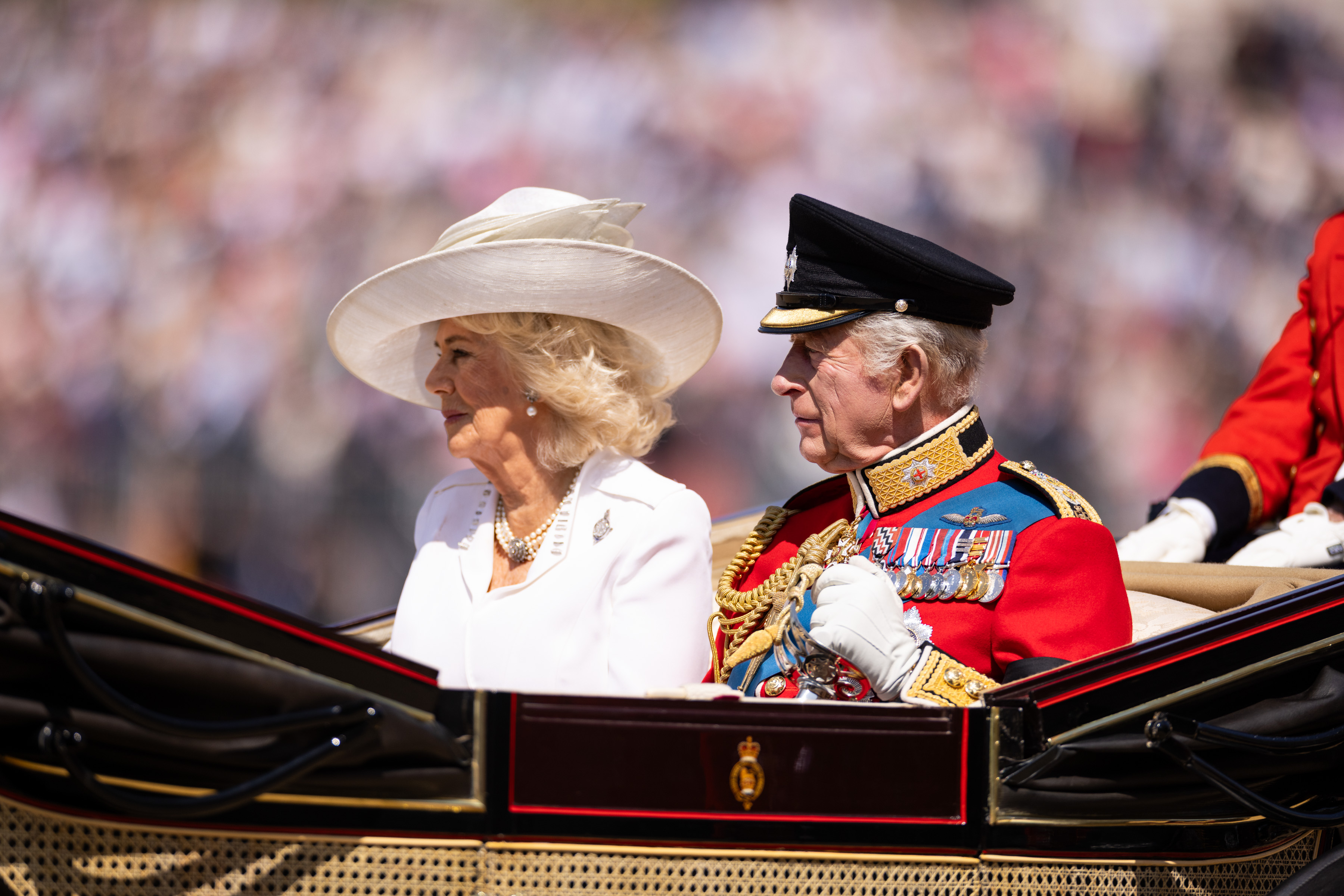 Her Majesty The Queen wearing a white dress and hat sits to the right of His Majesty The King who is wearing British Army ceremonial attire.