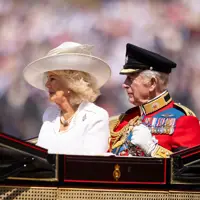 Her Majesty The Queen wearing a white dress and hat sits to the right of His Majesty The King who is wearing British Army ceremonial attire.