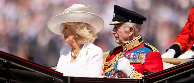 Her Majesty The Queen wearing a white dress and hat sits to the right of His Majesty The King who is wearing British Army ceremonial attire.