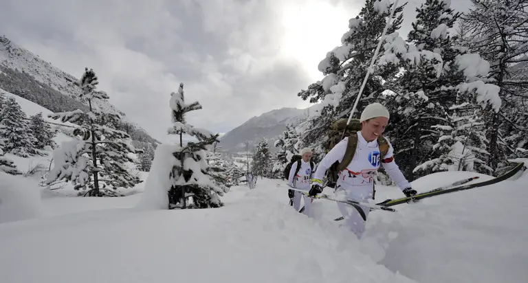 Female soldiers ski team walking through knee high snow while holding their ski's. Deep snow, trees and mountains can be seen in the background.