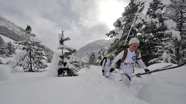Female soldiers ski team walking through knee high snow while holding their ski's. Deep snow, trees and mountains can be seen in the background.