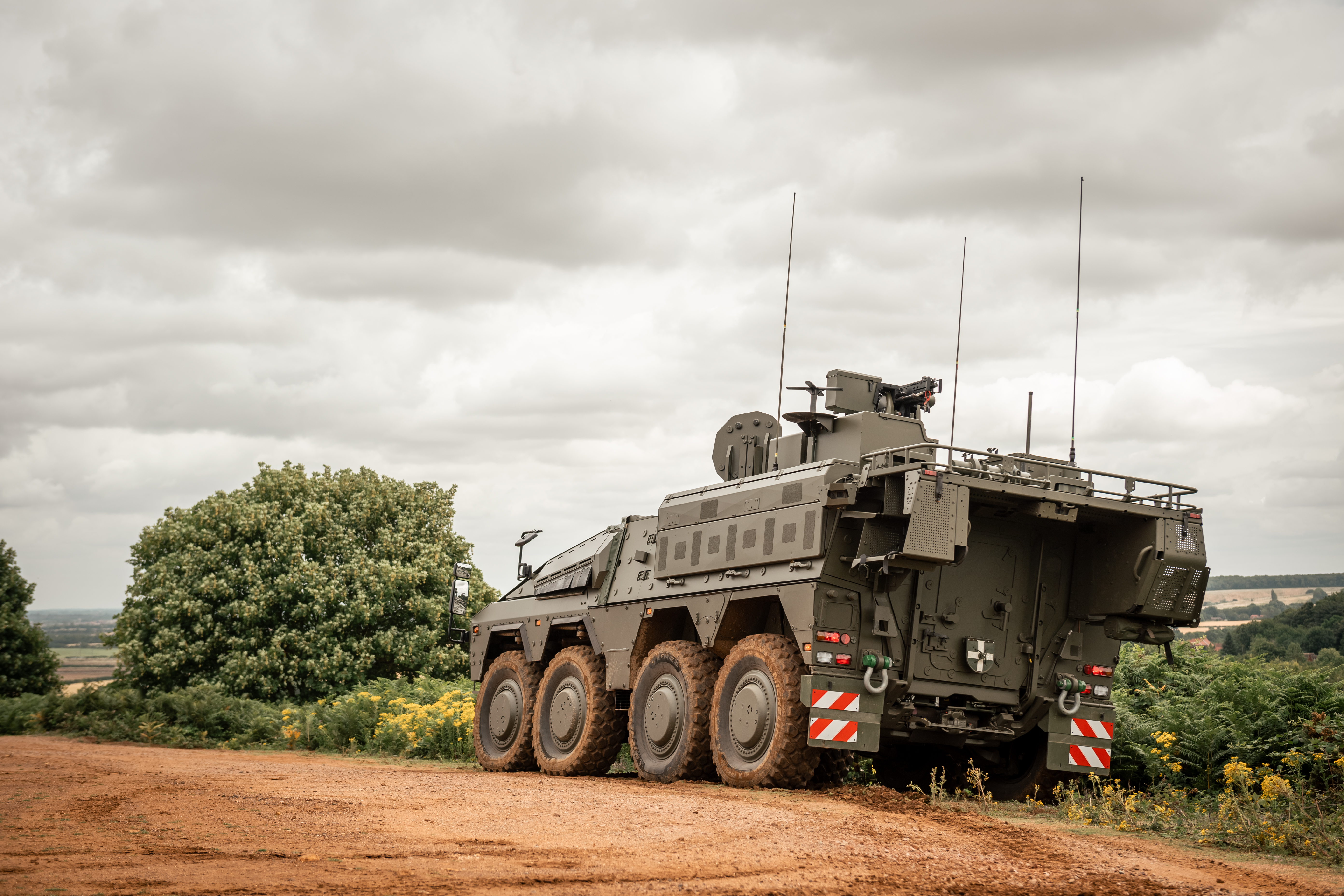 An armoured military vehicle with eight wheels is positioned on a dirt road. The background features green foliage.