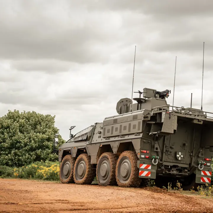 An armoured military vehicle with eight wheels is positioned on a dirt road. The background features green foliage.