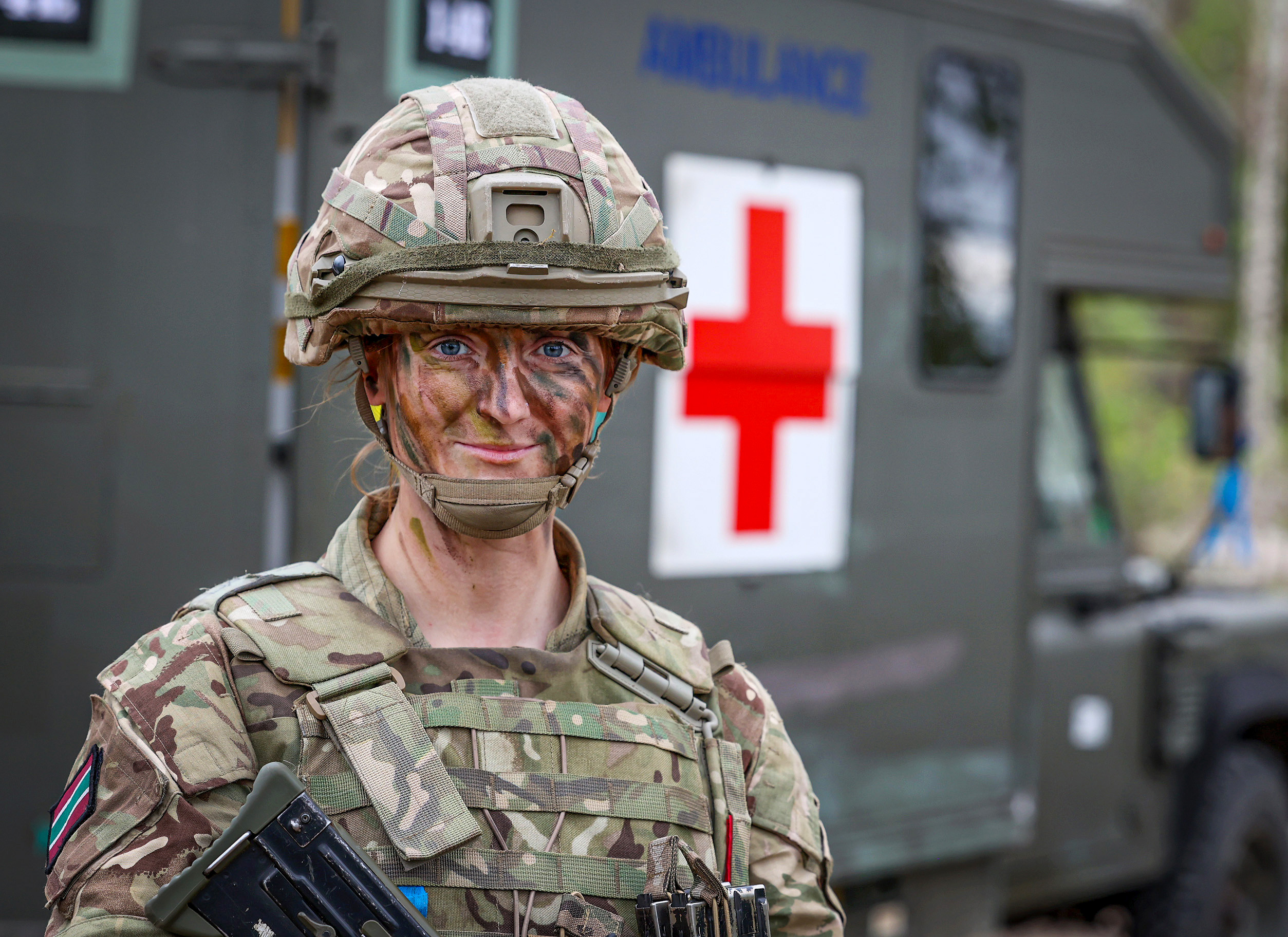 A soldier in camouflage gear and face paint stands confidently in front of a green military ambulance with a large red cross symbol. The scene conveys readiness.