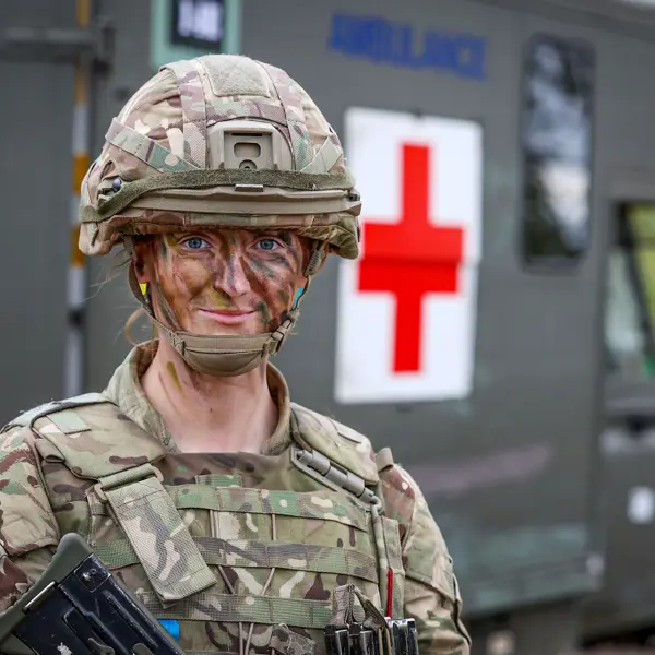 A soldier in camouflage gear and face paint stands confidently in front of a green military ambulance with a large red cross symbol. The scene conveys readiness.