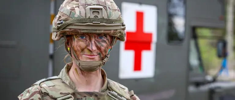 A soldier in camouflage gear and face paint stands confidently in front of a green military ambulance with a large red cross symbol. The scene conveys readiness.