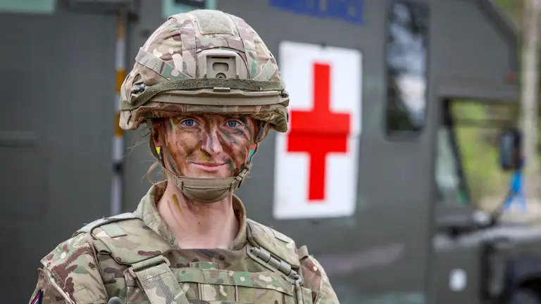 A soldier in camouflage gear and face paint stands confidently in front of a green military ambulance with a large red cross symbol. The scene conveys readiness.