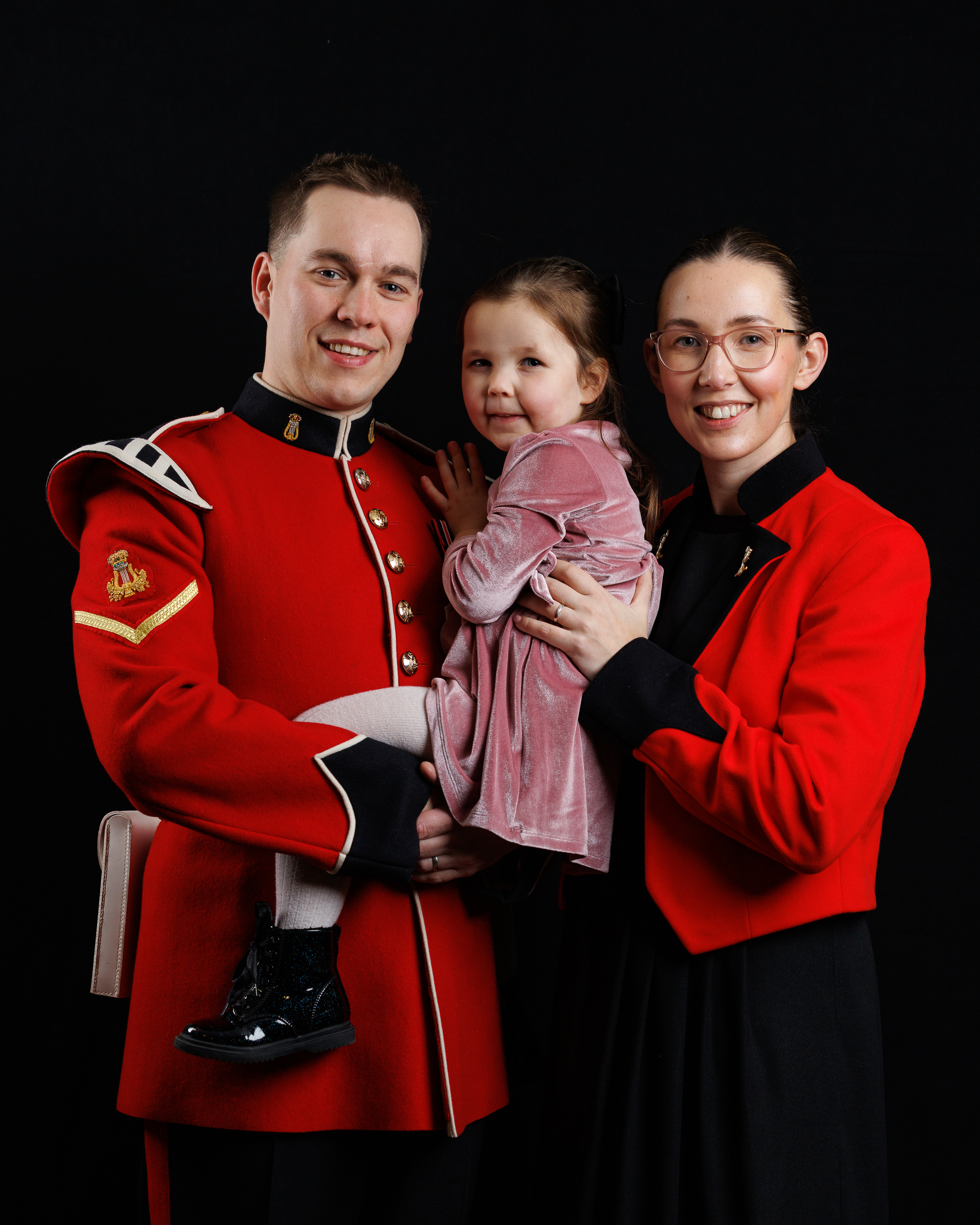 A man in a red military uniform holding a young girl in a pink dress, standing next to a woman wearing the same attire.