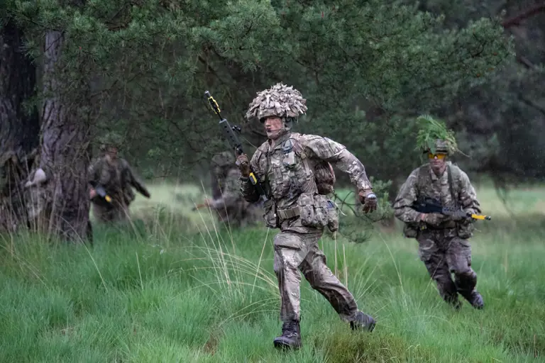 Officer cadets in camouflage uniform seen running through long grass holding their rifles