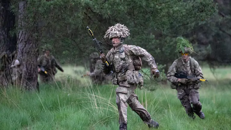 Officer cadets in camouflage uniform seen running through long grass holding their rifles